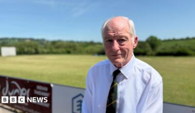 Tony Evans, a man with white hair, sits with his back to the cricket match, wearing a white shirt and green and yellow Purnell Cricket Club tie.