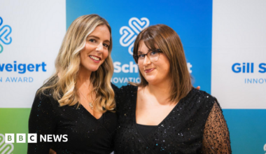 Leah and Florence are both wearing black, shimmery dresses and are stood in front of a blue, white and green advertisement board. Leah has long, wavy blonde hair and Florence has bobbed brown hair and glasses.