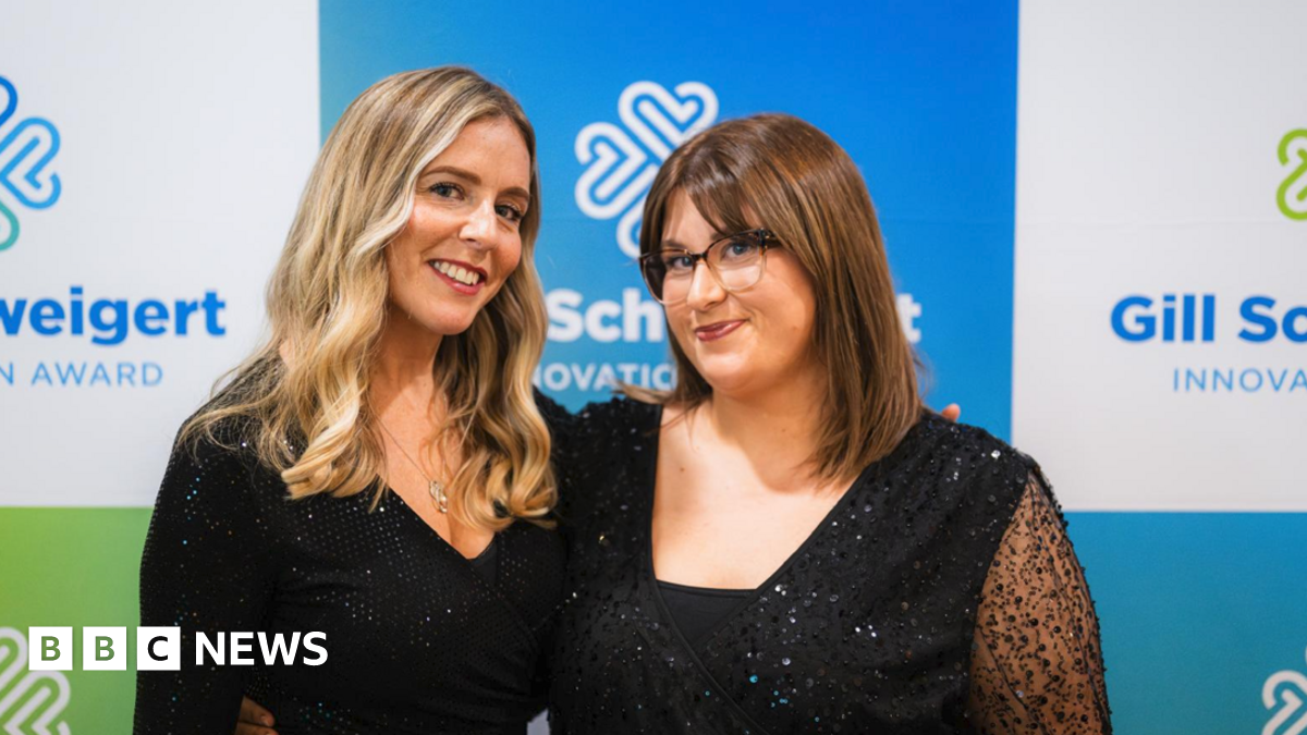 Leah and Florence are both wearing black, shimmery dresses and are stood in front of a blue, white and green advertisement board. Leah has long, wavy blonde hair and Florence has bobbed brown hair and glasses.