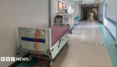 An empty portable hospital bed is at the side of a long empty hospital corridor. There are portable stands behind it with monitors and equipment.