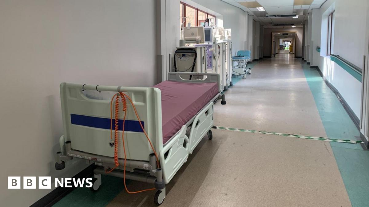 An empty portable hospital bed is at the side of a long empty hospital corridor. There are portable stands behind it with monitors and equipment.