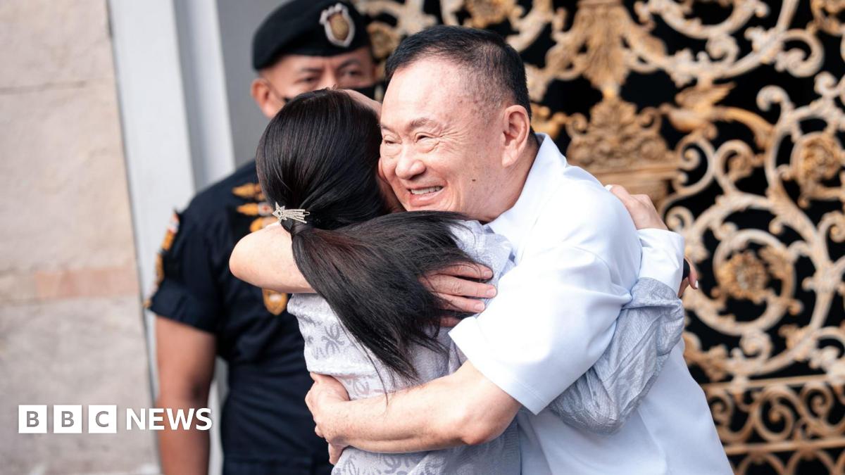 Thaksin, smiling and holding his hands in a prayer pose, stands among a sea of supporters