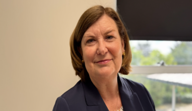 Wendy Reid has short straight brown hair. She is wearing a dark blue suit jacket over a patterned top. She is standing in an office in front of a window.
