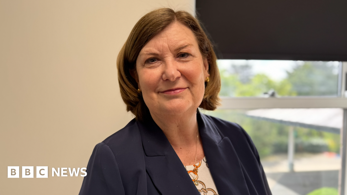 Wendy Reid has short straight brown hair. She is wearing a dark blue suit jacket over a patterned top. She is standing in an office in front of a window.
