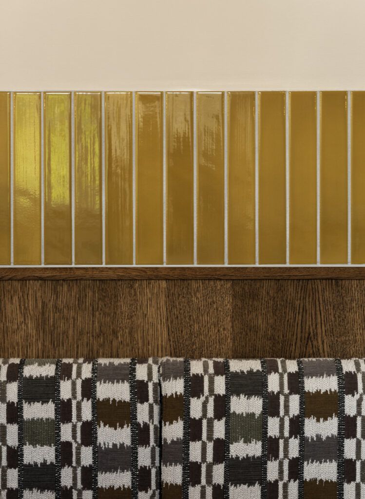 A sophisticated, retro-inspired restaurant with wood paneled walls, dark green upholstered banquettes, glazed golden tiles placed atop patterned black and white fabrics, and sculptural lights.