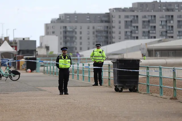 Two police officers seen beside a cordon on a pier, with a black waste bin visible alongside a teal railing.