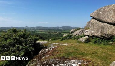A picture of Helman Tor in Cornwall. A wide countryside landscape with lots of green fields with grass land and hills in the background. There are large rocks to the left under a clear blue sky.