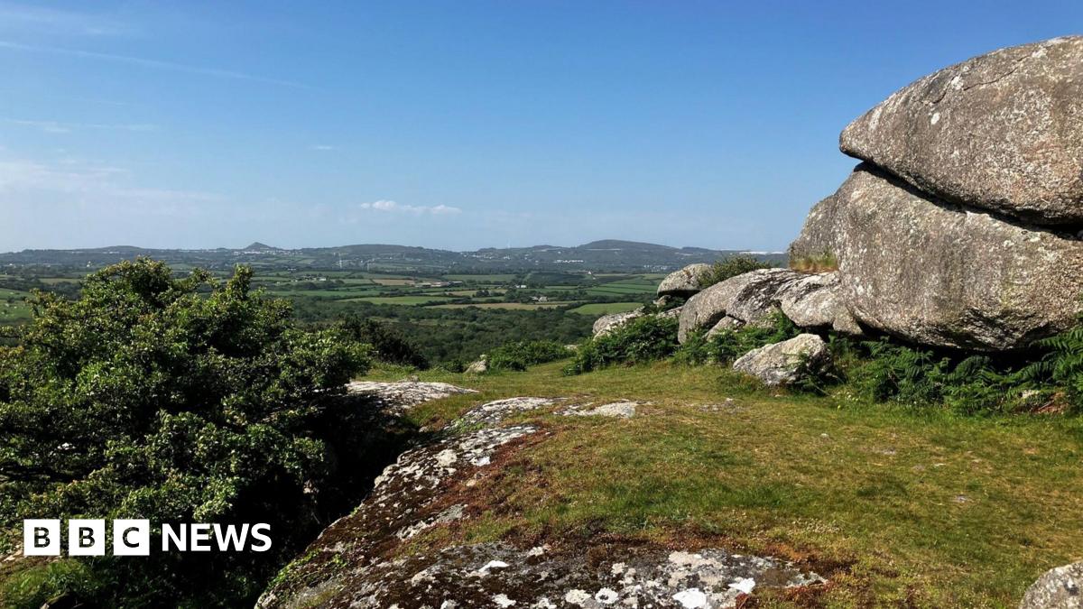 A picture of Helman Tor in Cornwall. A wide countryside landscape with lots of green fields with grass land and hills in the background. There are large rocks to the left under a clear blue sky.