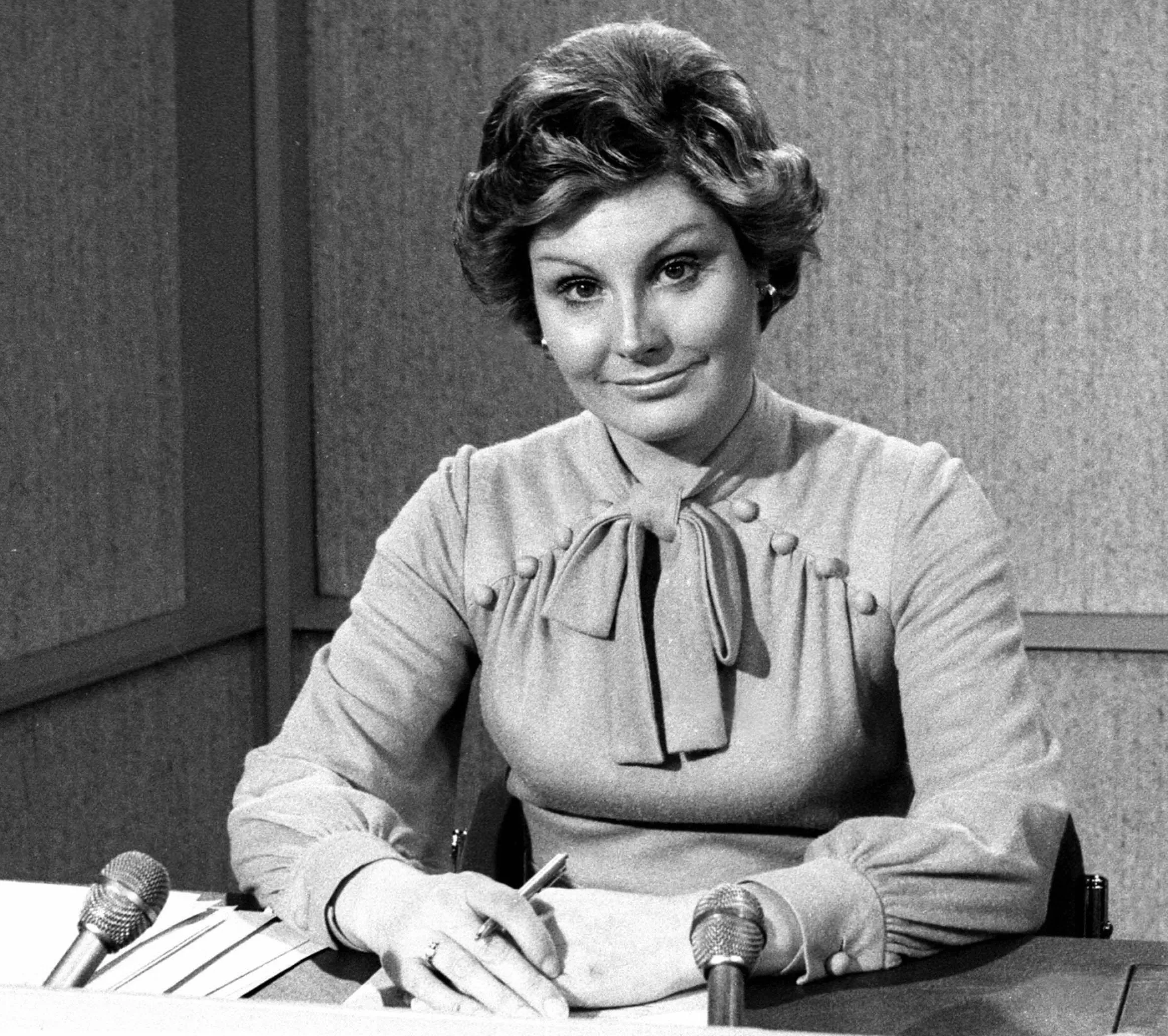 Newsreader Angela Rippon smiling at a desk with two microphones.