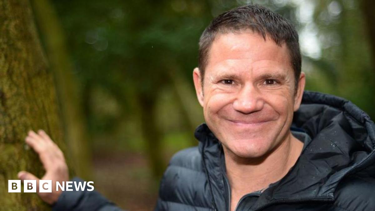 A man with brown hair is smiling at the camera. He is wearing a black puffer jacket and has one hand on a mossy tree beside him