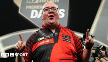 Stephen Bunting celebrates, pointing with both index fingers and pulling a celebratory facial pose as he wears a black and red darts shirt