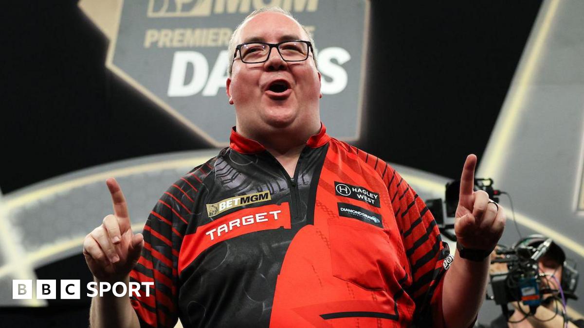 Stephen Bunting celebrates, pointing with both index fingers and pulling a celebratory facial pose as he wears a black and red darts shirt