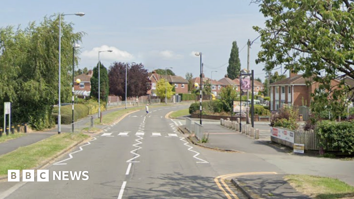 A suburban street lined by estate housing with trees either side and a pedestrian crossing visible
