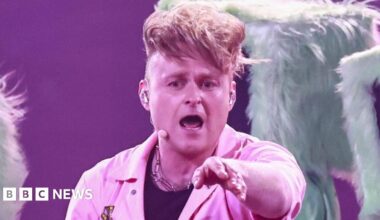 Sam Battle aka Look Mum No Computer performing on stage during Eurovision dress rehearsals in a pink jumpsuit pointing towards the camera, with three dancers behind him wearing fur-lined computer monitors with red lips over their heads