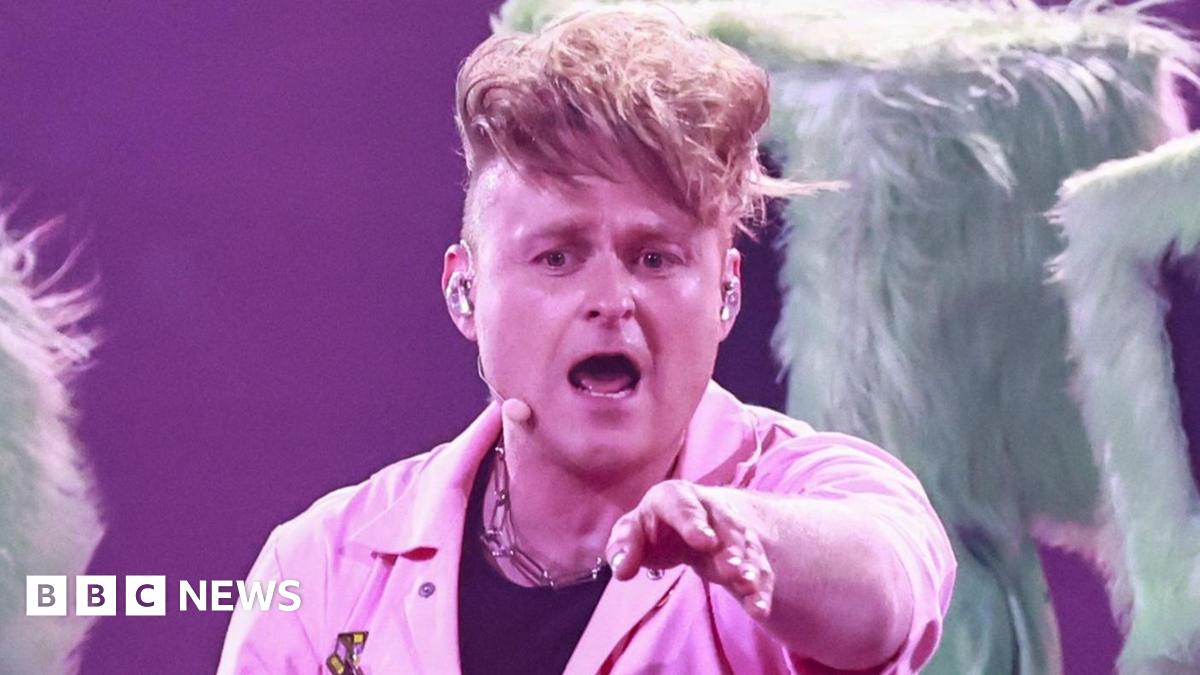 Sam Battle aka Look Mum No Computer performing on stage during Eurovision dress rehearsals in a pink jumpsuit pointing towards the camera, with three dancers behind him wearing fur-lined computer monitors with red lips over their heads