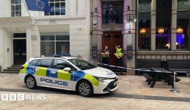 A police car is parked diagonally across a kerb outside a bar in a city street. There are black wooden benches outside obstructed by police tape, and two officers standing outside the ornate wooden doors of the building.