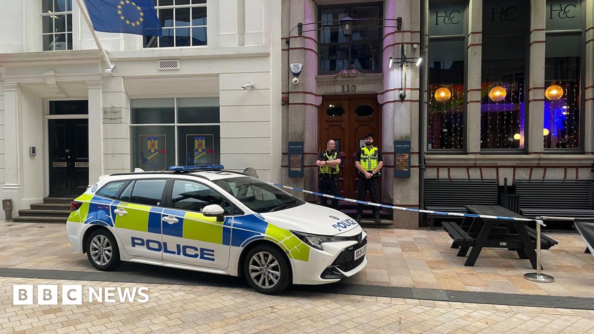 A police car is parked diagonally across a kerb outside a bar in a city street. There are black wooden benches outside obstructed by police tape, and two officers standing outside the ornate wooden doors of the building.