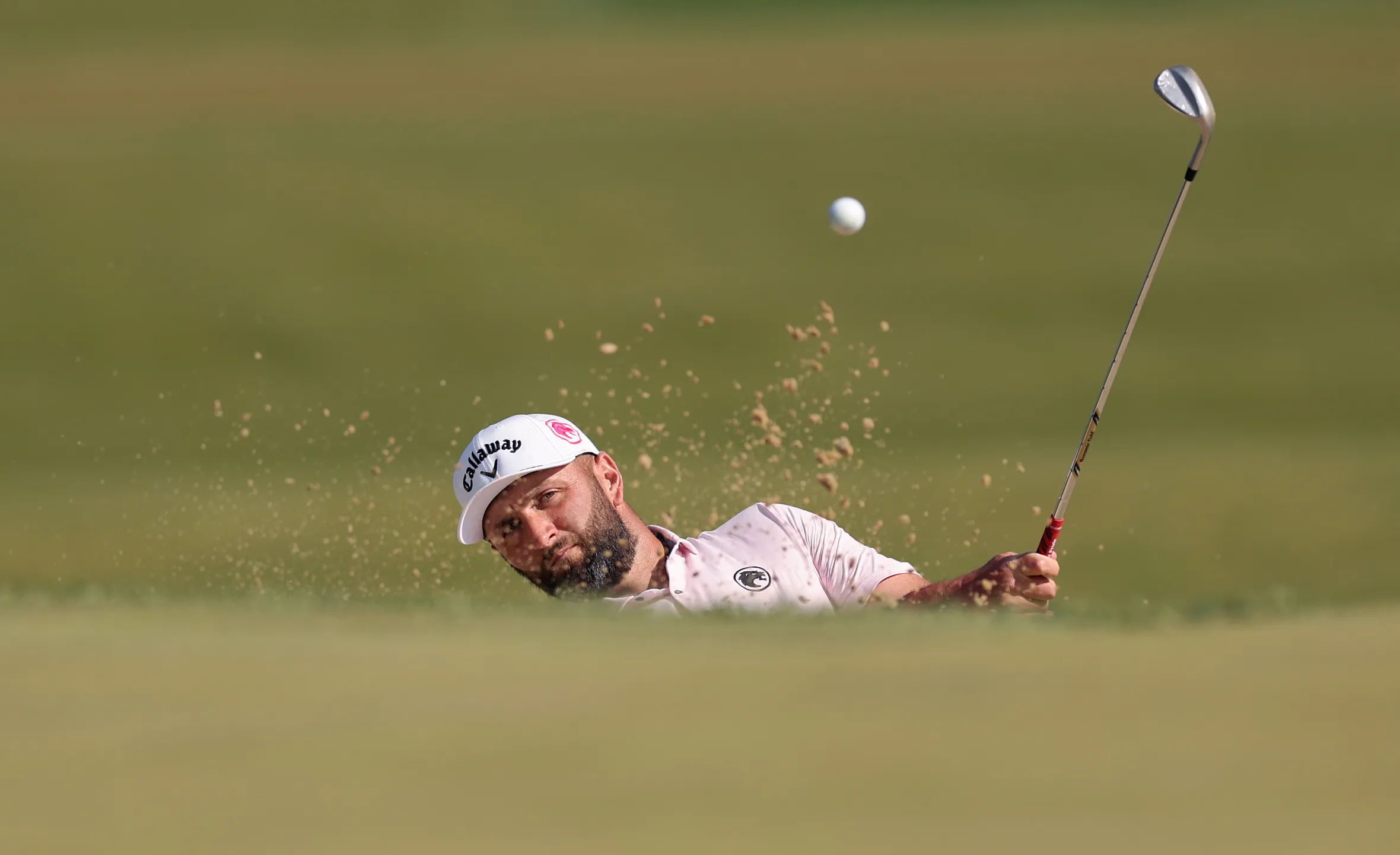 Jon Rahm hitting a golf ball out of a greenside bunker.