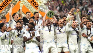 The Aston Villa team, made up of about 20 men, all wearing white uniforms with blue accents, are grouped together cheering. In the centre one of them is holding a large trophy above his head
