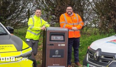 A picture of two people stood either side of the talking bin. It is brown in colour and there is a vehicle either side of the bin.