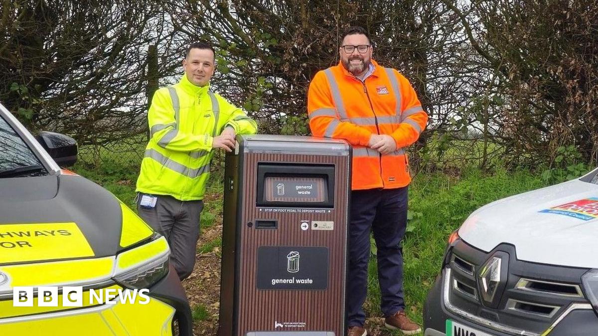 A picture of two people stood either side of the talking bin. It is brown in colour and there is a vehicle either side of the bin.