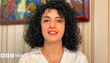A woman with curly black hair dressed in a white collared shirt and red lipstick, smiles to the camera.