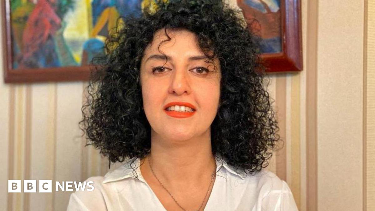 A woman with curly black hair dressed in a white collared shirt and red lipstick, smiles to the camera.