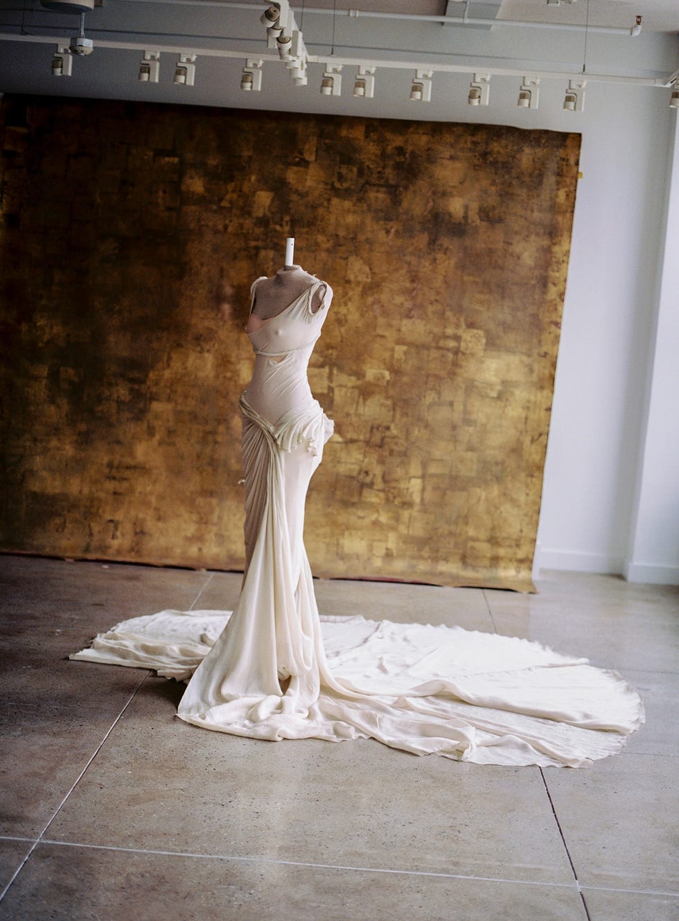Elegant draped gown displayed on a dress form against a textured golden backdrop.