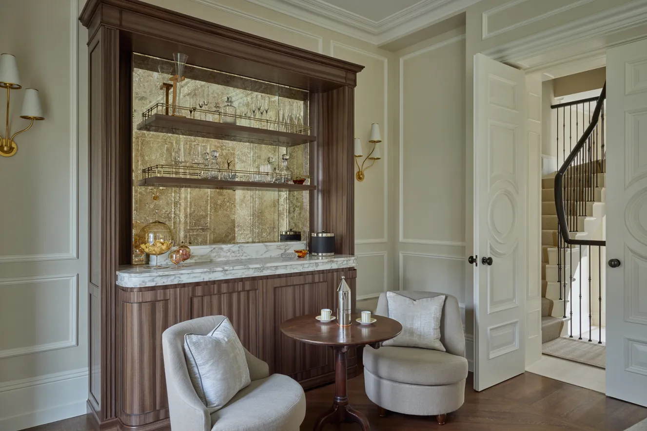 A bespoke home bar by Katharine Pooley with Calacatta Oro marble top, two light-colored armchairs, and a small round table with coffee cups.