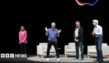 Apple CEO Tim Cook on a dark stage with a neon Apple logo behind him. He is wearing a blue polo sweater, black jeans and white sneakers and gesturing his hands, while three other people on stage look at him.