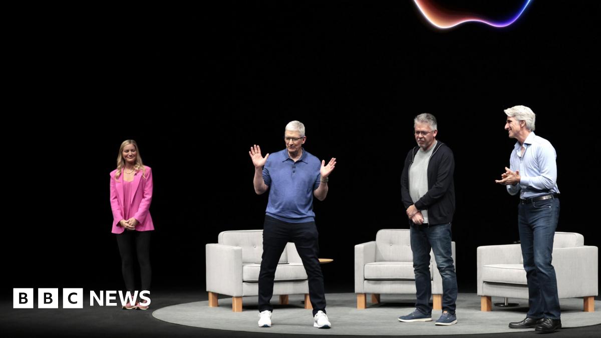 Apple CEO Tim Cook on a dark stage with a neon Apple logo behind him. He is wearing a blue polo sweater, black jeans and white sneakers and gesturing his hands, while three other people on stage look at him.