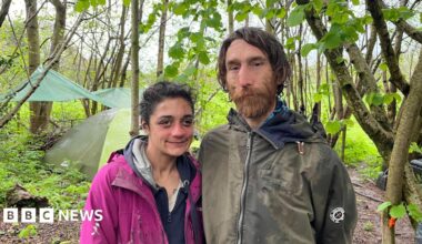 A man and woman stand in a wood in front of a green tent and tarpaulin tied between trees. The man has long brown hair, a brown beard and a green coat. The woman has black hair tied back, a navy hoodie and a pink coat.