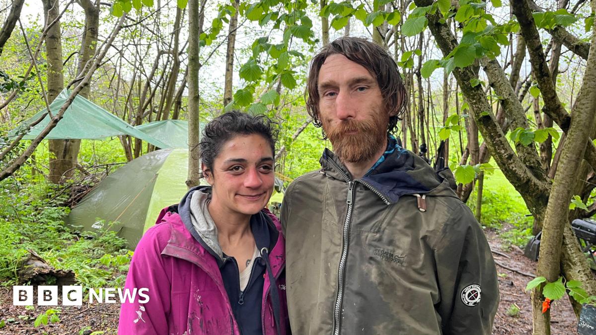 A man and woman stand in a wood in front of a green tent and tarpaulin tied between trees. The man has long brown hair, a brown beard and a green coat. The woman has black hair tied back, a navy hoodie and a pink coat.