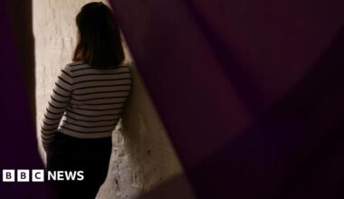 A woman in a white top with black stripes leans against the lit area of a white wall.