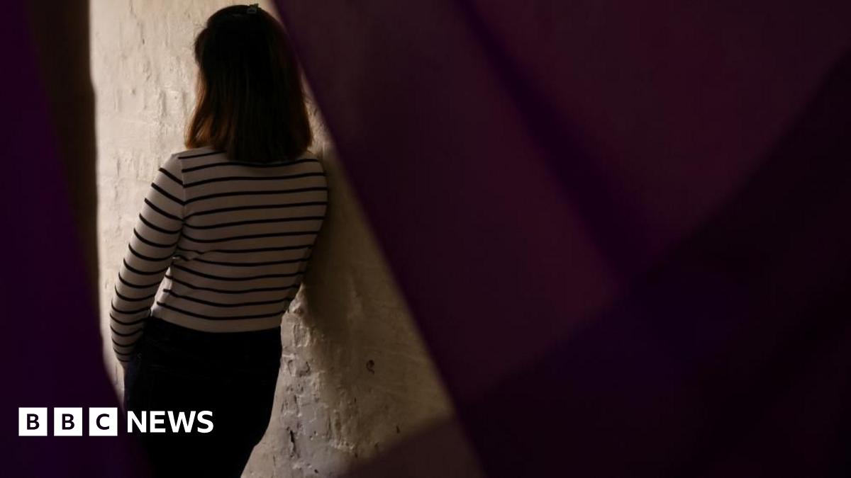 A woman in a white top with black stripes leans against the lit area of a white wall.