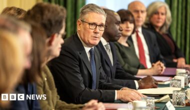 Sir Keir Starmer addresses members of the cabinet. He is looking towards the camera and is wearing glasses and a dark suit and tie with a white shirt. Some members of the cabinet are looking away from the camera and some are not in focus. In front of them are papers, drinks glasses and mugs.