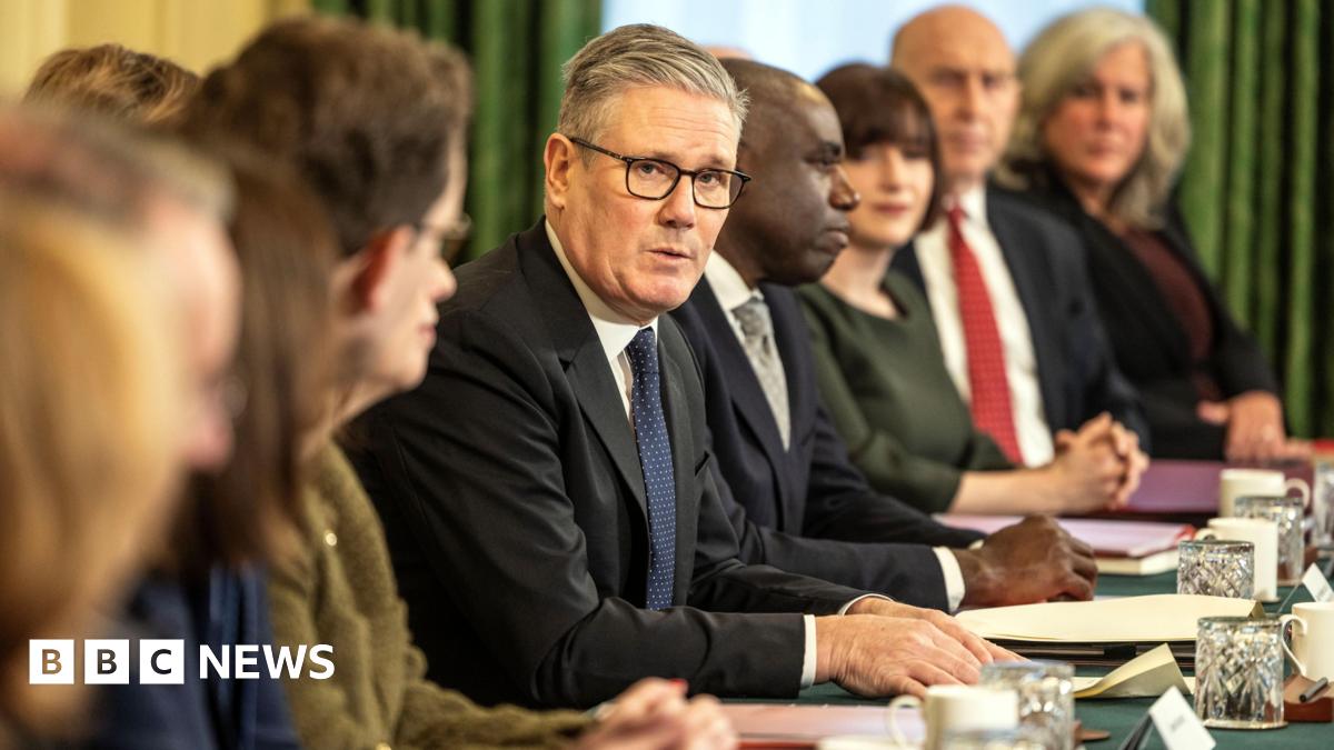 Sir Keir Starmer addresses members of the cabinet. He is looking towards the camera and is wearing glasses and a dark suit and tie with a white shirt. Some members of the cabinet are looking away from the camera and some are not in focus. In front of them are papers, drinks glasses and mugs.