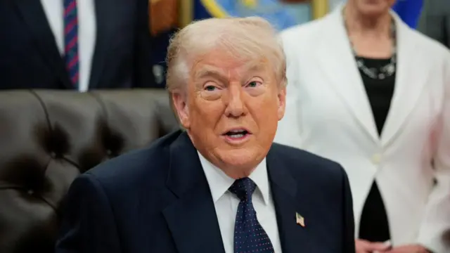 Donald Trump speaks while sitting in the Oval Office. He is wearing a dark suit and navy tie with red and white dots.