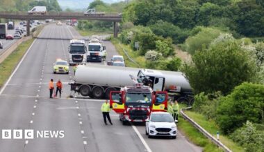 An empty motorway with a truck across the road and into the barrier and hedge. There is a fire truck and emergency services in high vis jackets.