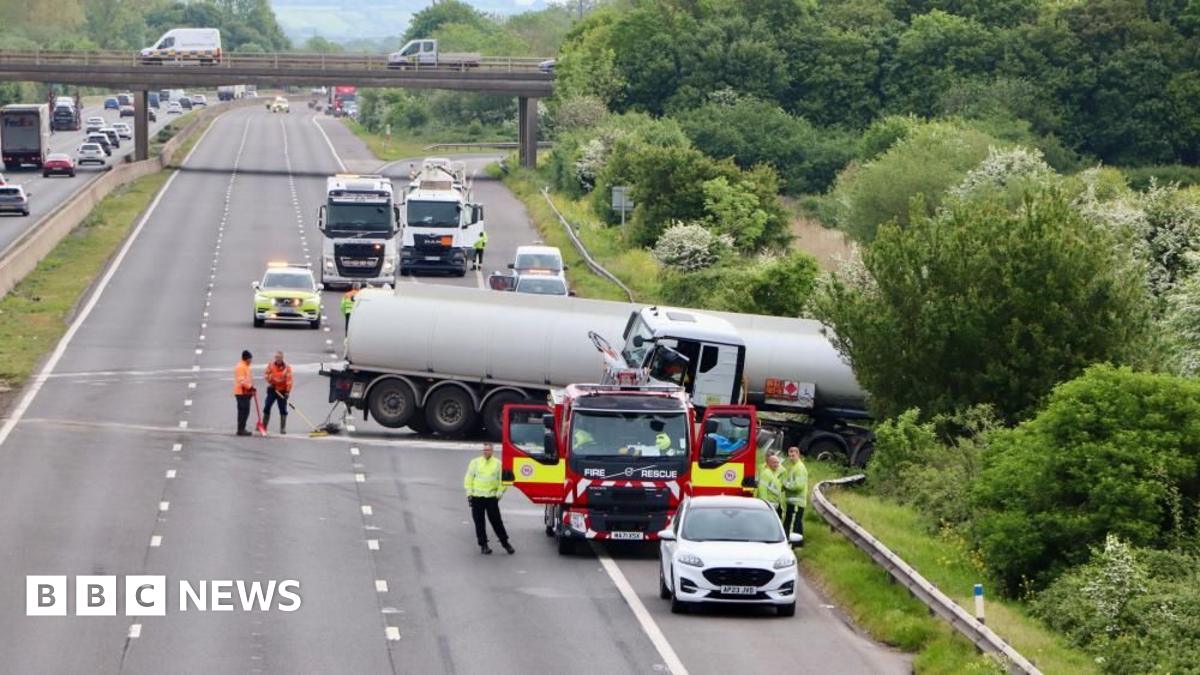 An empty motorway with a truck across the road and into the barrier and hedge. There is a fire truck and emergency services in high vis jackets.
