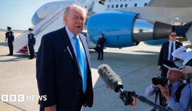 Trump wearing a suit and speaking to reporters, with a plane parked behind him.
