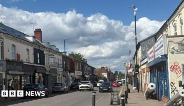 A main A road with shops lining either side and cars going back and forth. On the lampposts St George's flags are flying high
