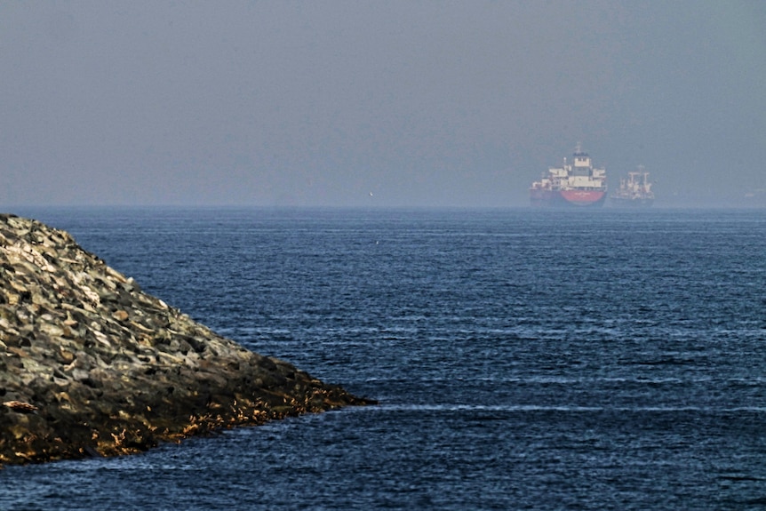 Two cargo ships can be seen in the distance on blue water, against blue skies. Rocky land is jutting out to sea on the left.