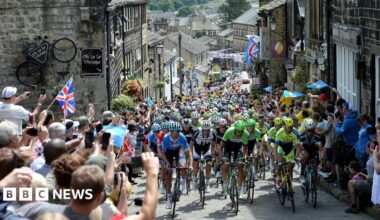 Cyclists climbing up a narrow street which has stone buildings on either side. Crowds line the street, a few holding Union Jack flags. There is bunting above the street further down the street which is the bottom of the road.