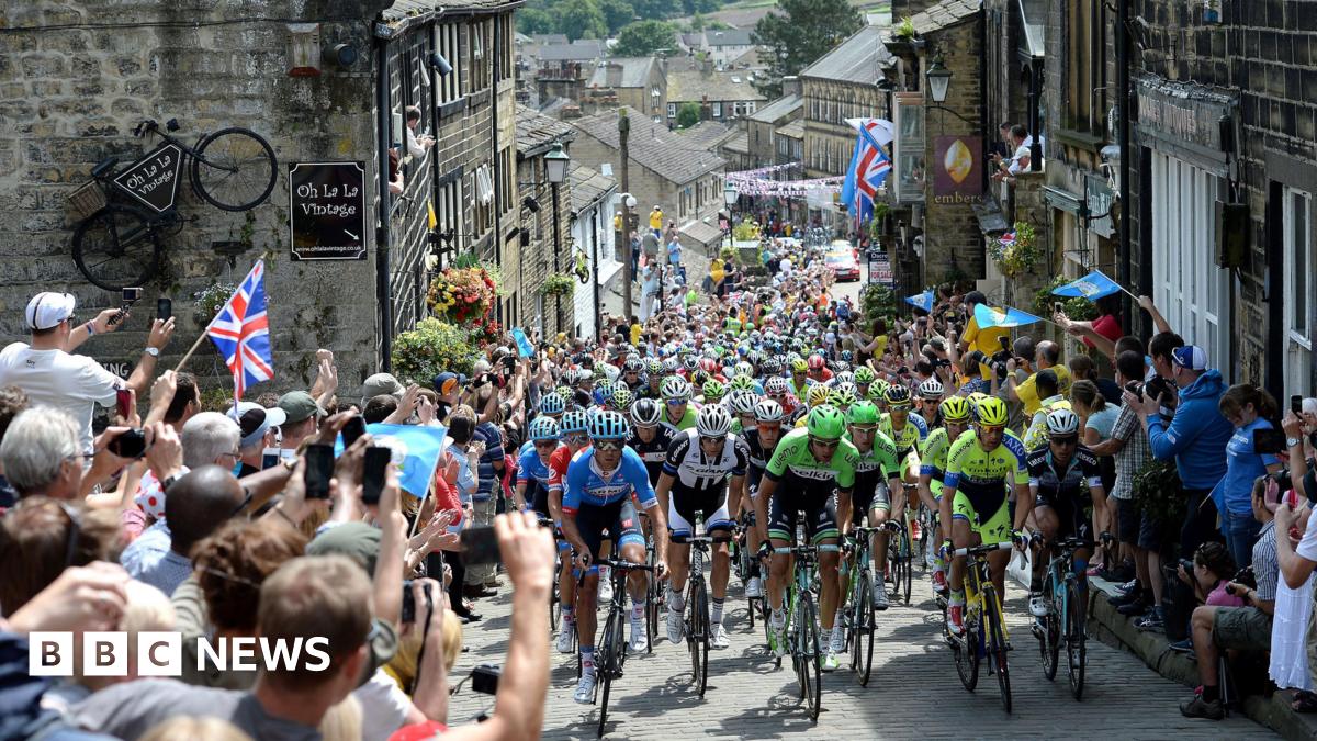 Cyclists climbing up a narrow street which has stone buildings on either side. Crowds line the street, a few holding Union Jack flags. There is bunting above the street further down the street which is the bottom of the road.