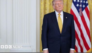 Donald Trump, in a dark navy suit and yellow tie, stands with his hands at his sides and a neutral expression. Behind him are gold curtains in the White House and an American flag.