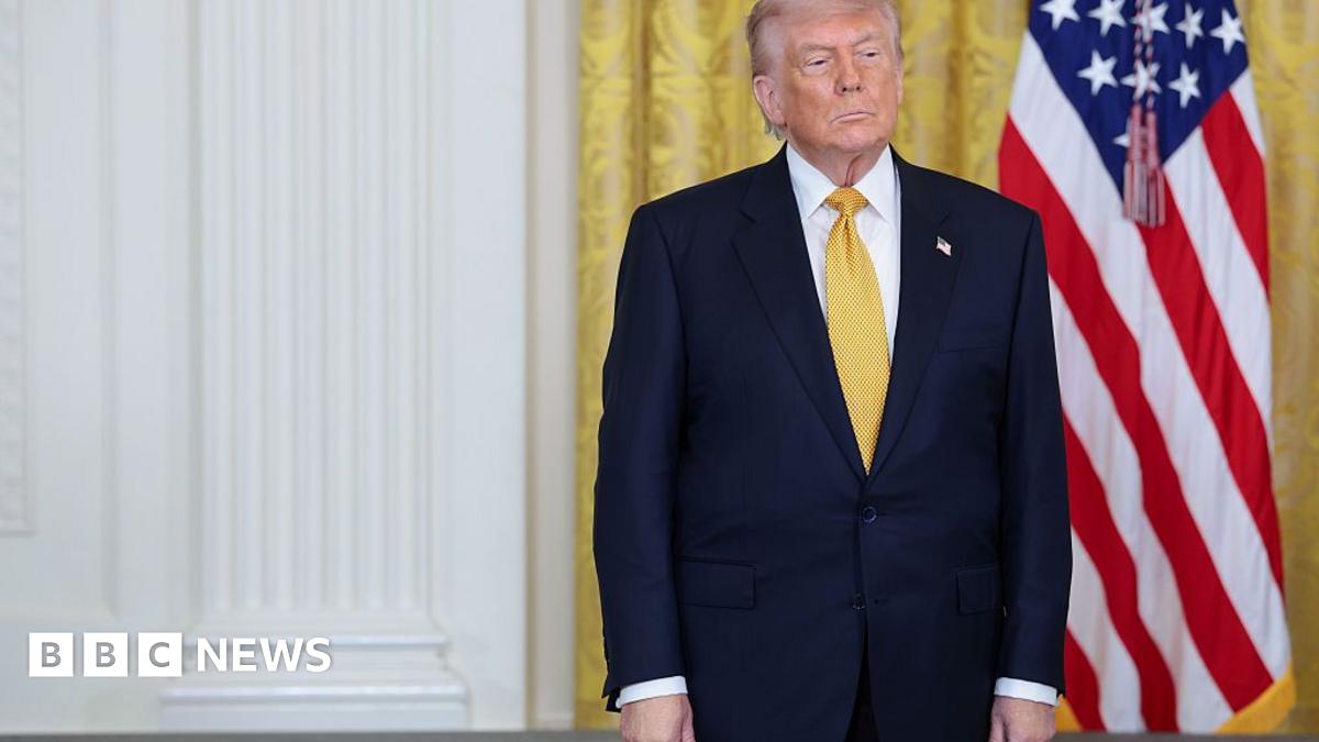 Donald Trump, in a dark navy suit and yellow tie, stands with his hands at his sides and a neutral expression. Behind him are gold curtains in the White House and an American flag.