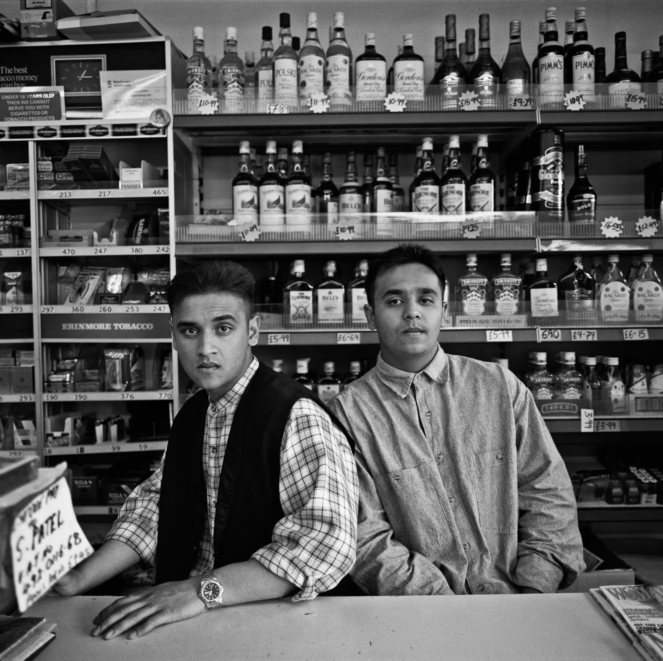 Sanjiv and Sandip, two young men, stand behind the counter of a store stocked with tobacco and alcohol.