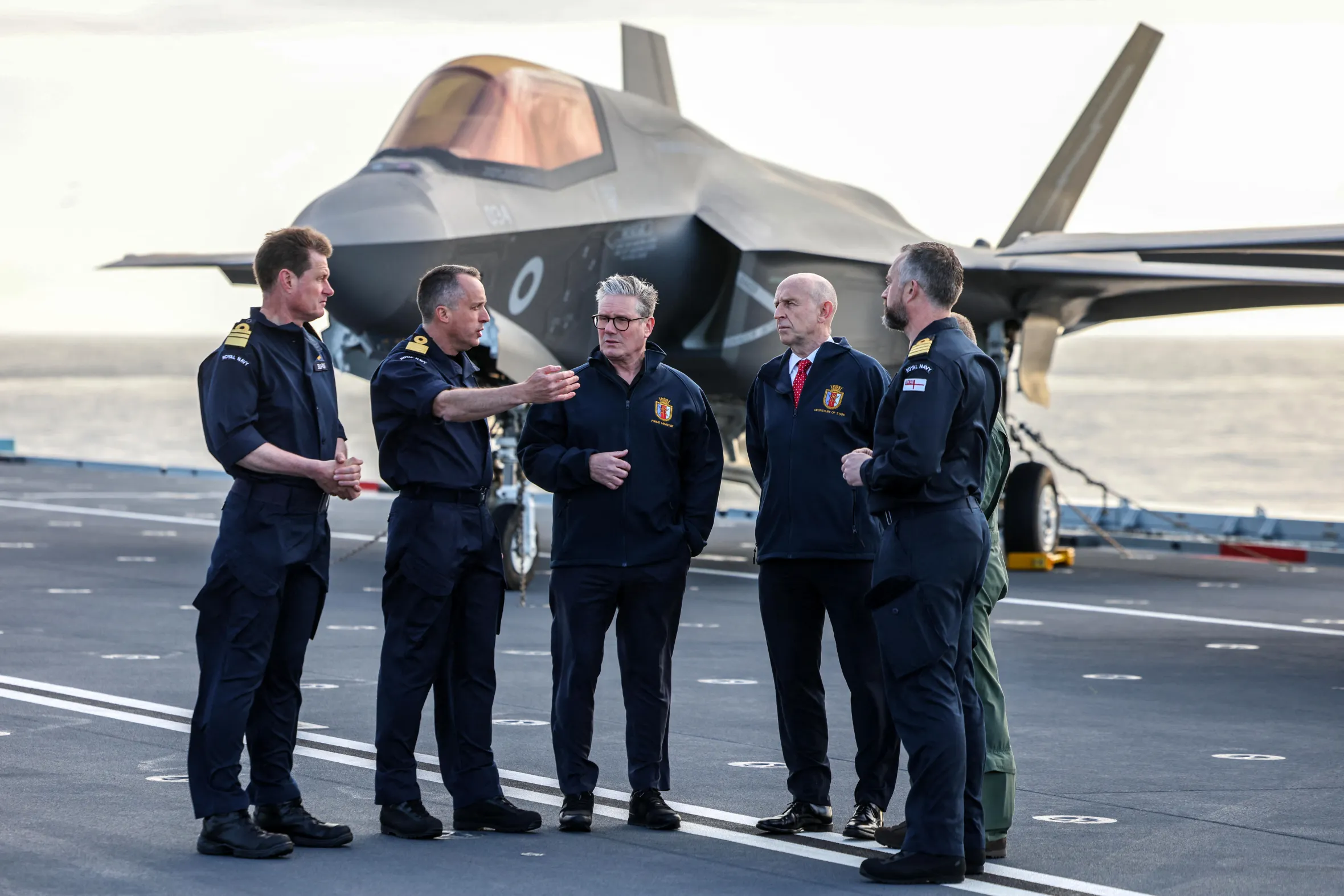 Prime Minister Keir Starmer and Defence Secretary John Healey on a ship deck with military personnel and an F-35B Lightning jet.