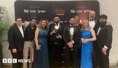 Nine men and women wearing various forms of formal evening dress pose for a picture at an awards ceremony. The man in the middle, in a dinner jacket and bow tie, is holding a black trophy. The group is standing in front of a black branded media wall featuring several sponsor names alongside the event name, The Signature Awards 2026.